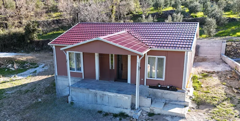 Kırmızı kiremit çatılı, önü kapalı verandalı tek katlı prefabrik evin yamaçtaki arsada havadan görünümü
